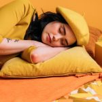 A fashionable woman in yellow with a beret sleeping next to a vintage alarm clock on a color-coordinated bed.